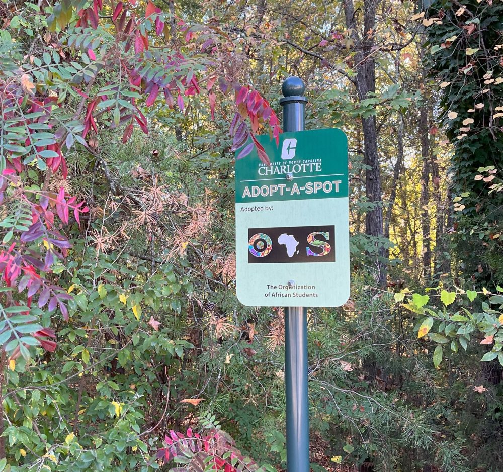 A sign that says Adopt a Spot with an organization's logo shown in front of fall foliage