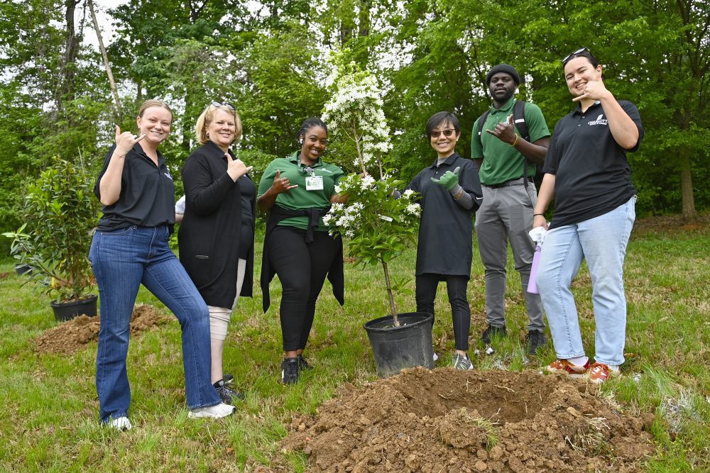 A group of UNC Charlotte staff pose doing the pickaxe sign with a tree