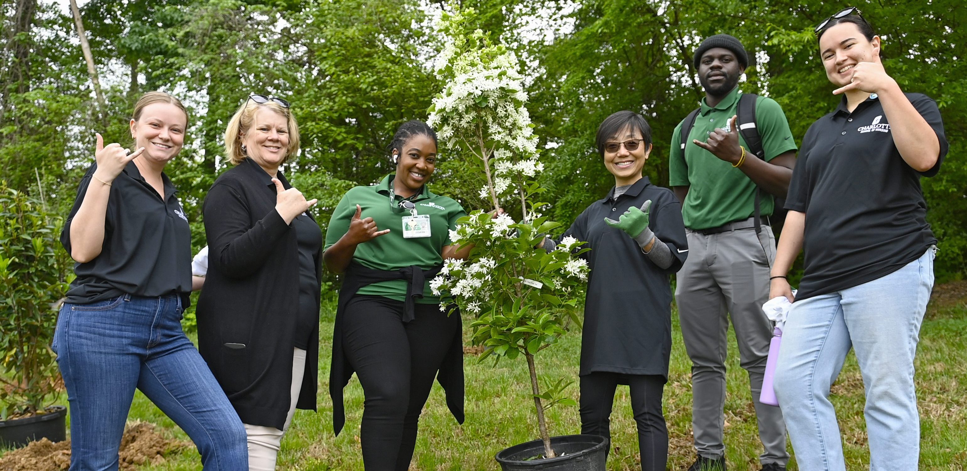 A group of UNC Charlotte staff pose doing the pickaxe sign with a tree