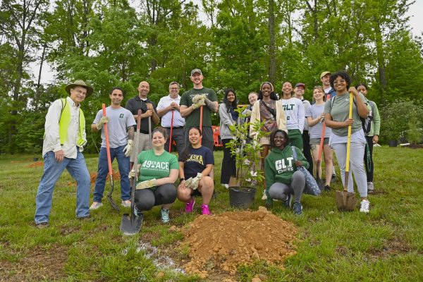 A group of students, staff, and faculty stand together in front of trees holding gardening tools